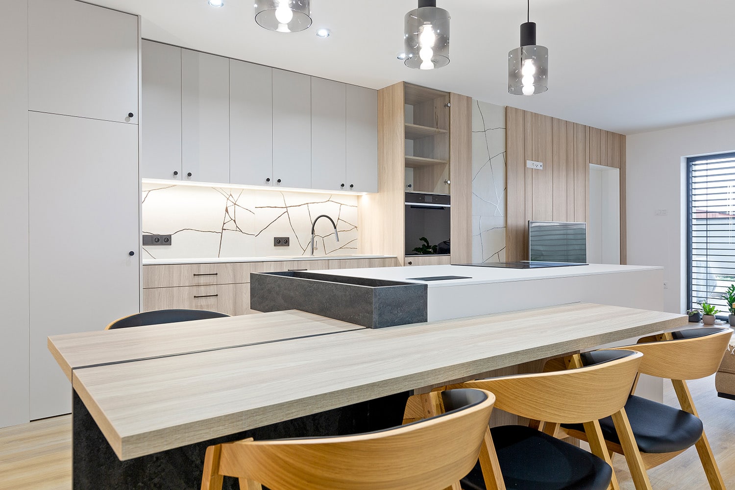moderni-kuchyne-s-jidelnim-prostorem Moderní kuchyně s ostrůvkovou kuchyní, jídelním stolem z přírodního dřeva a elegantními židlemi v propojení s obývacím pokojem.